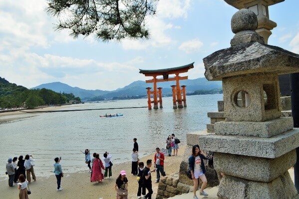 Tourists on the beach of Miyajima Island with an orange Torii gate floating in the background