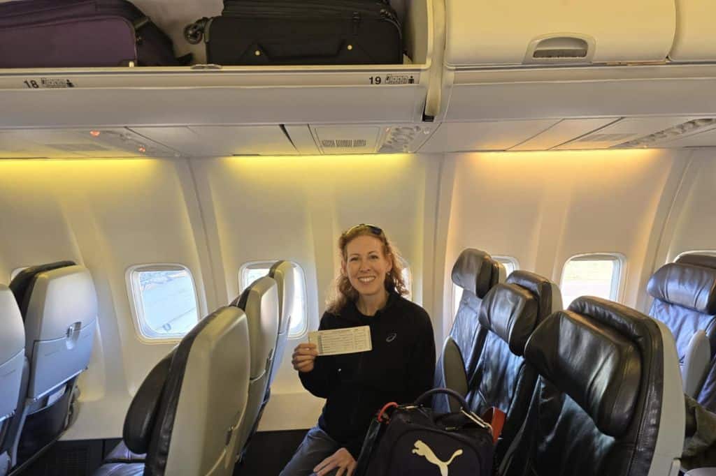 a woman sitting on a plane, holding her boarding pass. The overhead bin above her is open and shows two suitcases.