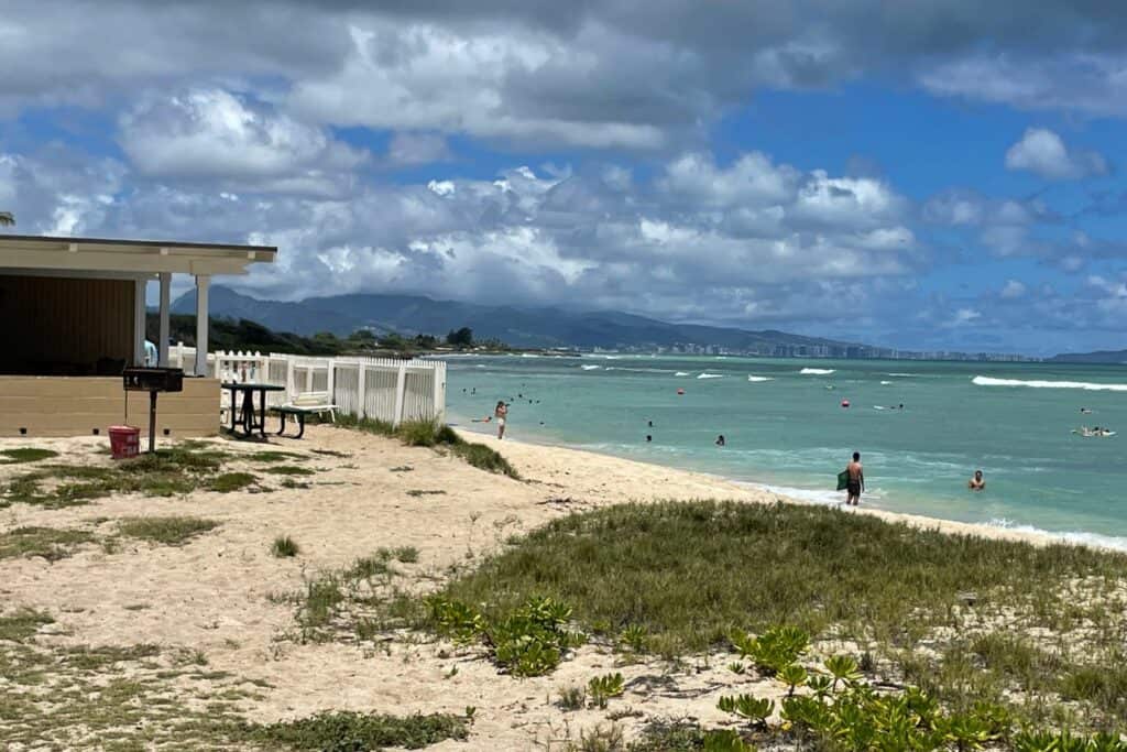 Cottage at Barbers Point on Oahu with white fence and picnic table overlooking a sandy beach and people swimming in the ocean.