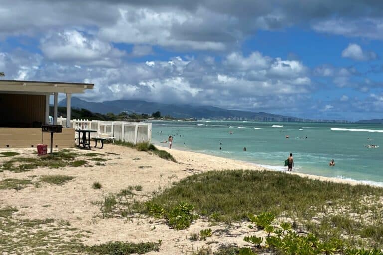 Cottage at Barbers Point on Oahu with white fence and picnic table overlooking a sandy beach and people swimming in the ocean.