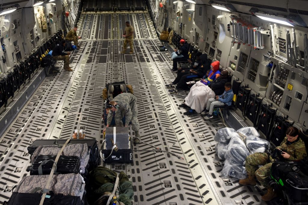 the inside of a military cargo plane with luggage strapped to the floor and passengers seated along the wall.