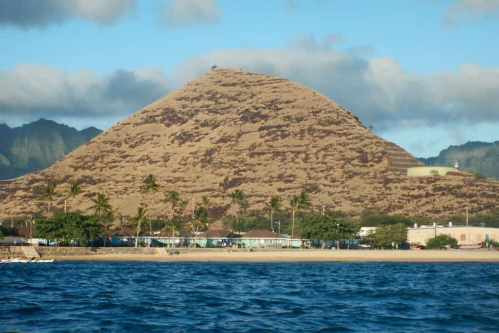 Beachfront view of Pililaau Army Recreation Center on Oahu, with small cottages and palm trees lining the shore, set against the backdrop of a large, brown, grass-covered mountain 