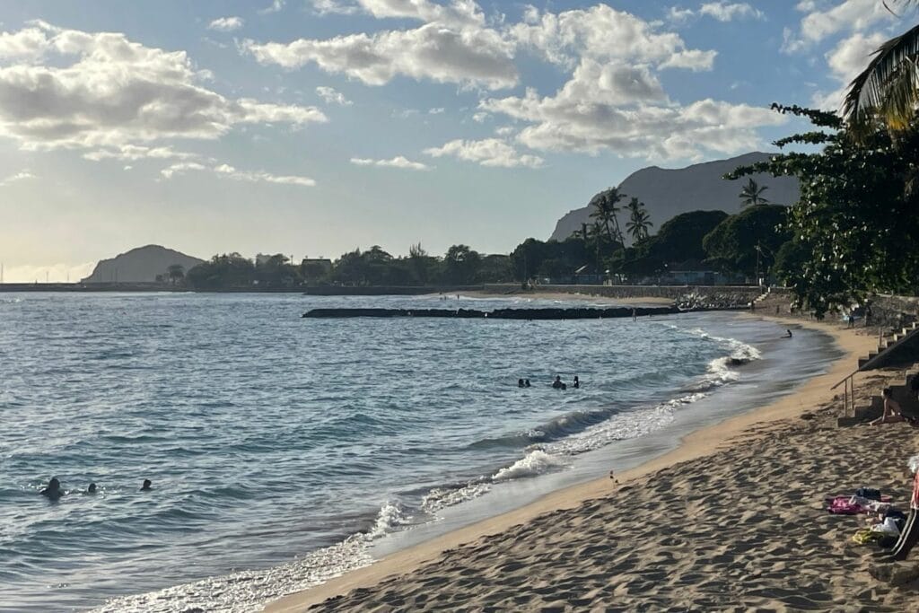 Gentle waves and swimmers along the sandy shoreline of Pokai Bay, framed by palm trees and mountains in the distance.