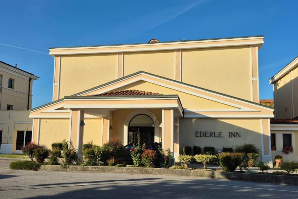 Exterior of the U.S. Army Health Center in Vicenza, a large cream-colored building with multiple towers, arched windows, and the facility name on a wall in front.