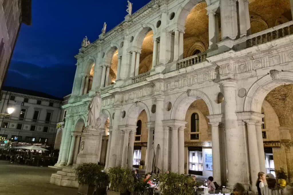 Night view of the Basilica Palladiana in Vicenza, with its white Renaissance arches and statues illuminated, and people sitting at restaurant tables in the square below