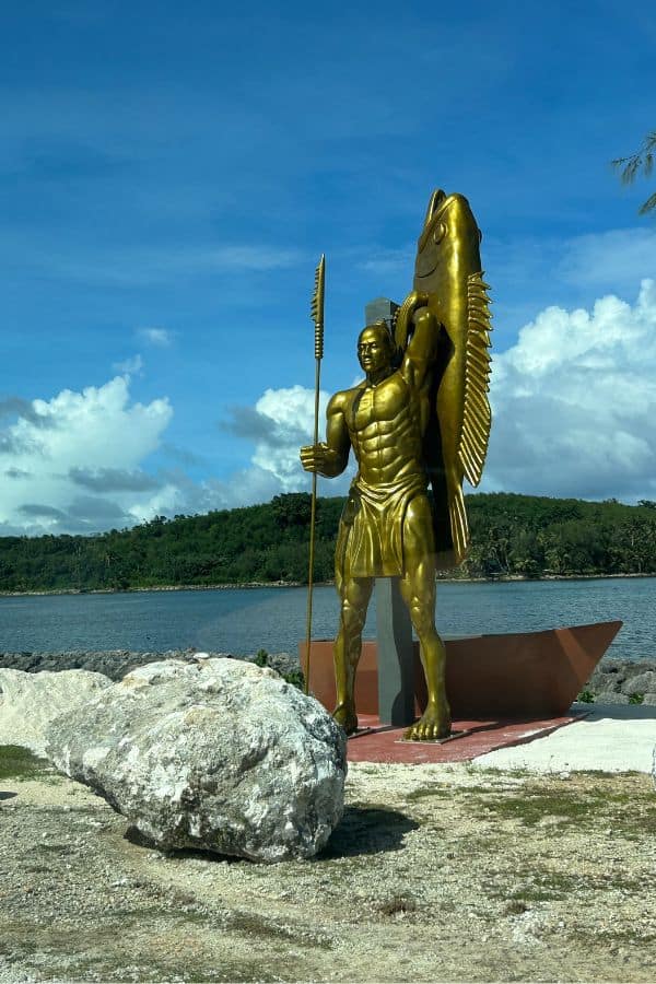 A golden statue of a Chamorro fisherman holding a spear and large fish, set against a blue sky
