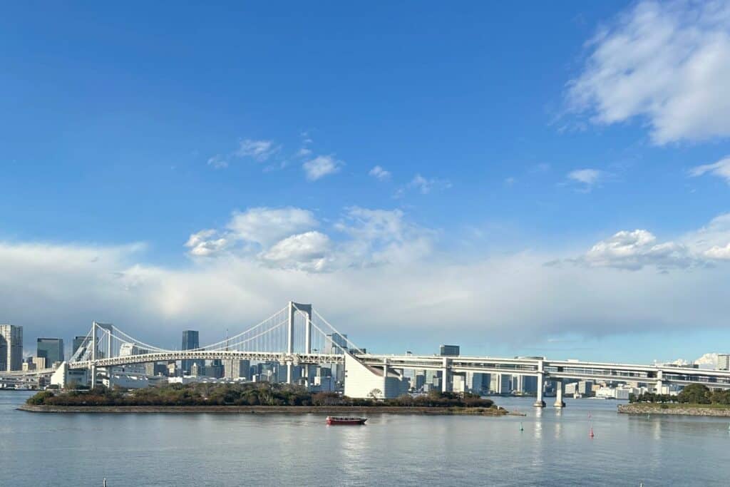 A suspension bridge spanning Tokyo Bay with skyline views and calm water in the foreground.