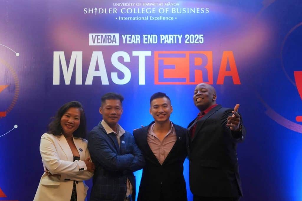 Markeiz and his classmates dressed in suits under a sign advertising the VEMBA Year End Party.