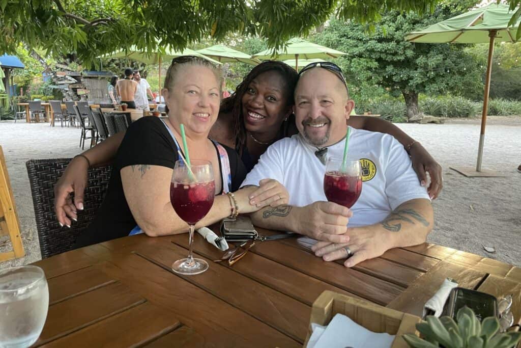 Isah smiles with two friends at an outdoor café in Costa Rica, with tropical trees and umbrellas in the background