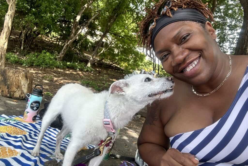 Isah smiles next to a small white dog on a beach blanket under the trees in Costa Rica.