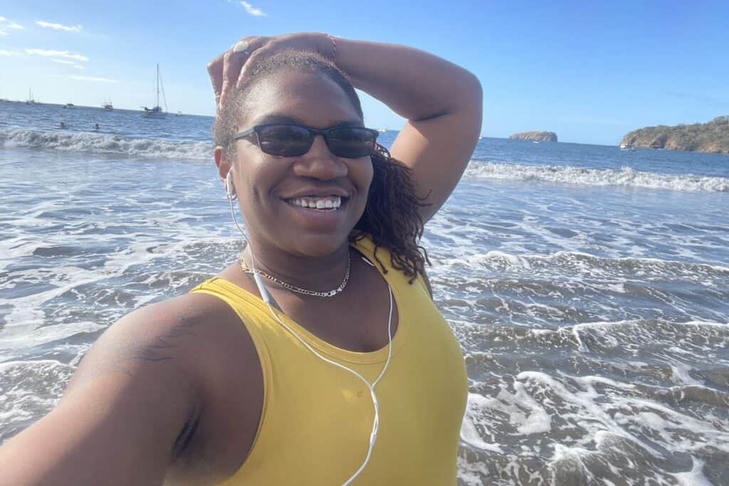 Isah takes a selfie on a beach in Guanacaste, Costa Rica, with boats and rocky coastline behind her.