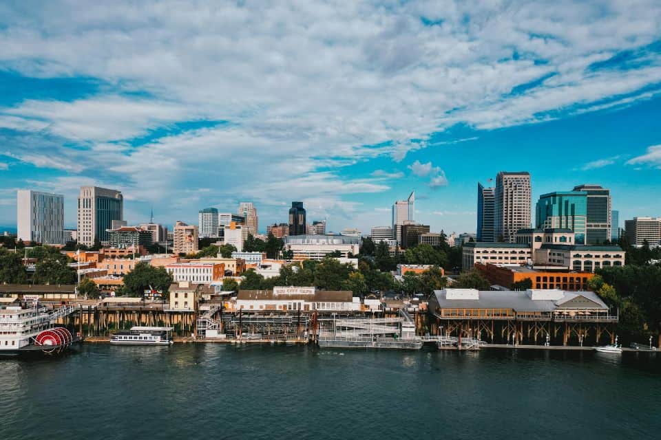Downtown Sacramento skyline along the river with buildings, boats, and a cloudy blue sky