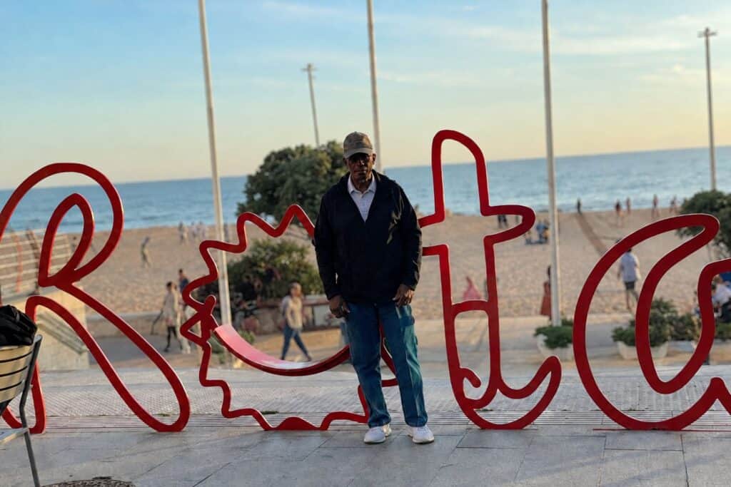 A man standing in front of a red sculpture that spells rota with the beach in the background