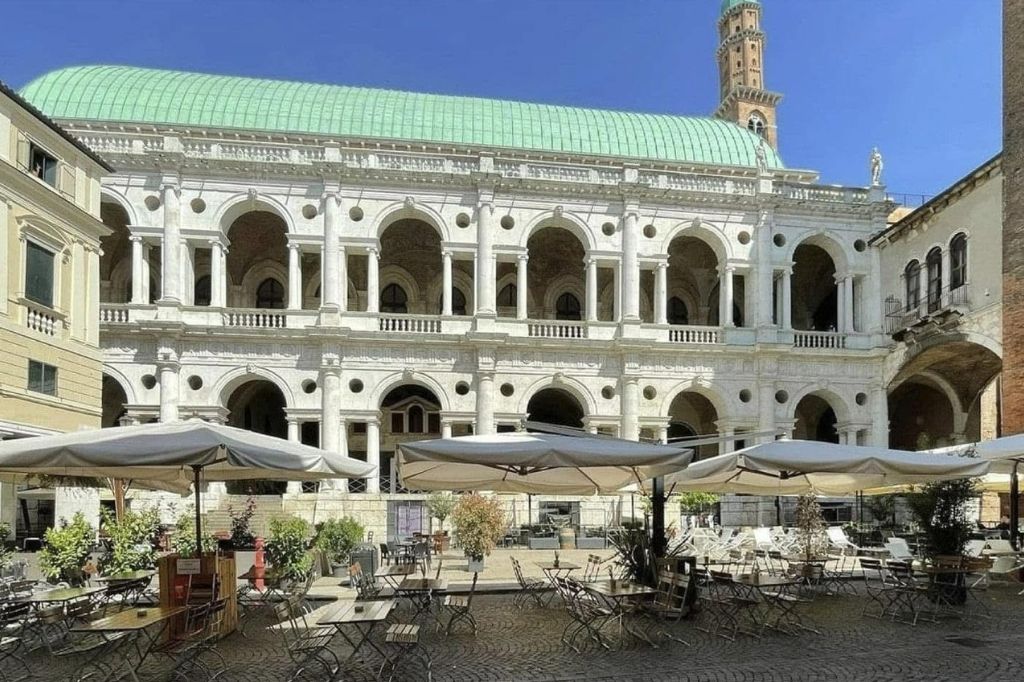 White Renaissance building with two levels of arches, a green roof, and café seating in front