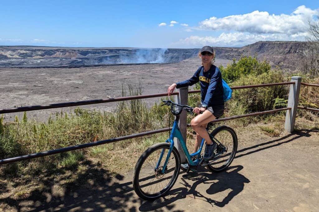 A woman on a bike in front of a crater with with smoke rising in the background