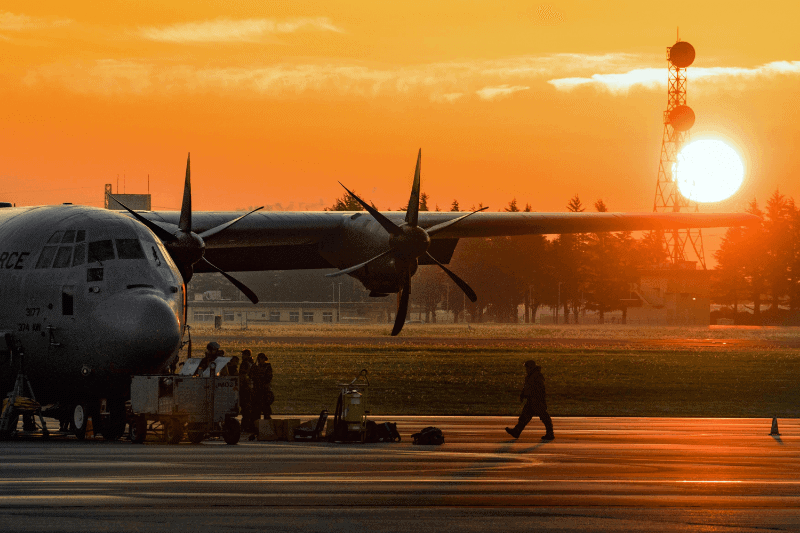 A military plane on the flight line with a bright sunrise in the background.