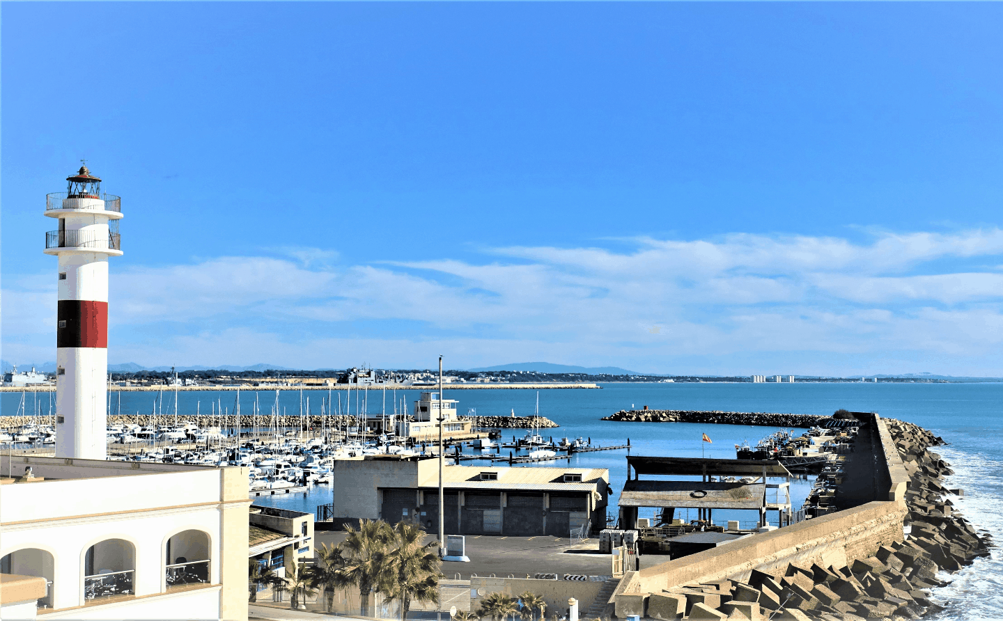 A red and white lighthouse next to Rota ferry terminal with boats in the harbor and Bay of Cadiz in the background