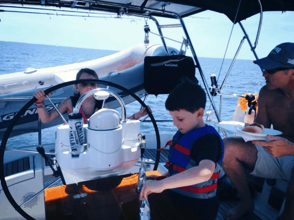 Two young children wearing life jackets and steering the boat, supervised by their dad.