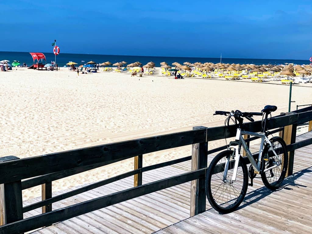 A bicycle parked along a wooden boardwalk with a beach full of umbrellas in the background