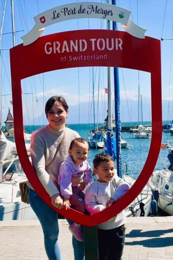 A woman and two young children under a sign for the Port de Morges with boats in the background