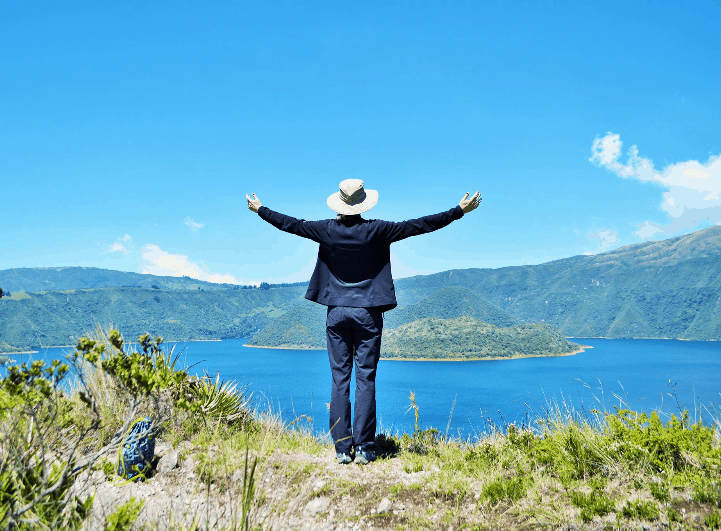 A woman standing with her back to the camera, arms spread to the sides, with a mountain lake in the background.