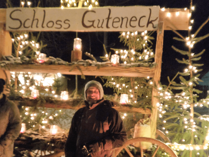 A man in a winter hat standing in front of trees with Christmas lights and a sign in German