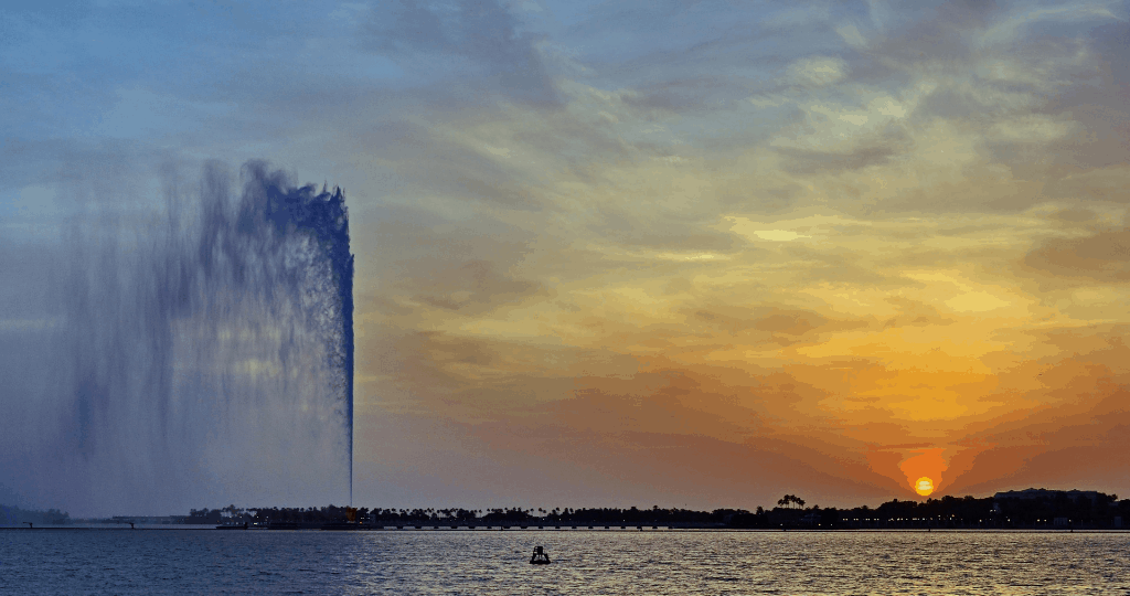 Picture of a tall fountain in front of the sea with a sunset in the background